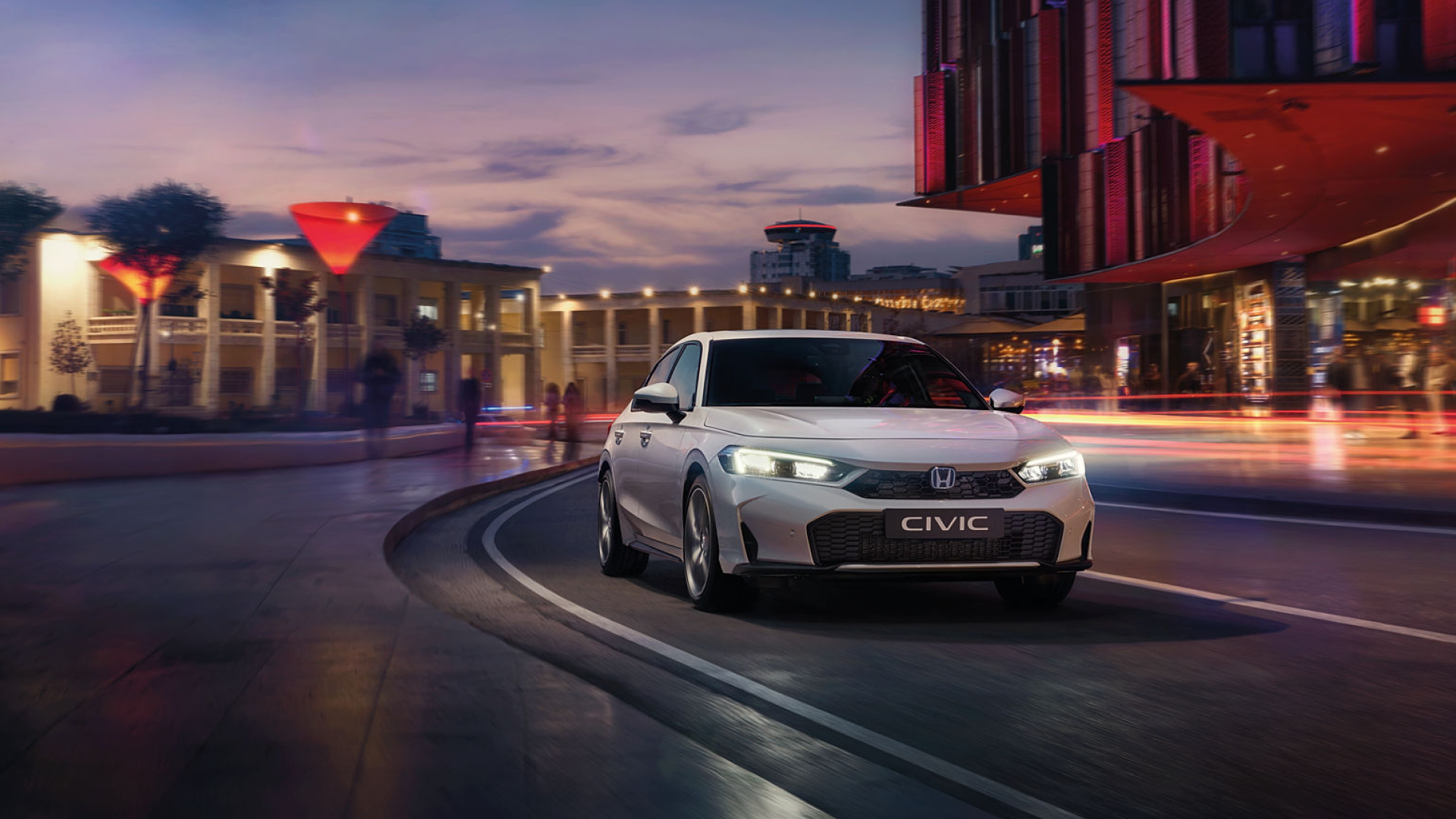 A white Honda Civic drives on a curved road at dusk in a city setting. The car's headlights are on, illuminating the path ahead. The background shows modern buildings with colorful lighting and a twilight sky.